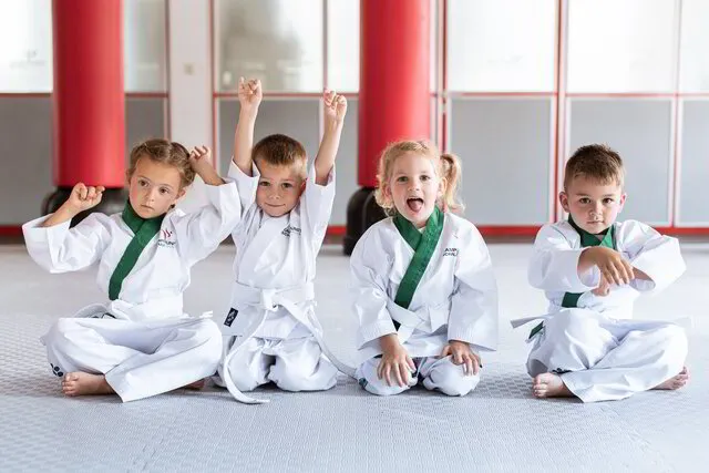 Vier kleine Karate Kinder sitzen im Dojo in Karate Anzügen auf dem Boden und albern herum, es sind zwei Mädchen und zwei Jungs im Alter von 3-5 Jahren der Kampfkunstschule Geiger, sie haben Spaß
