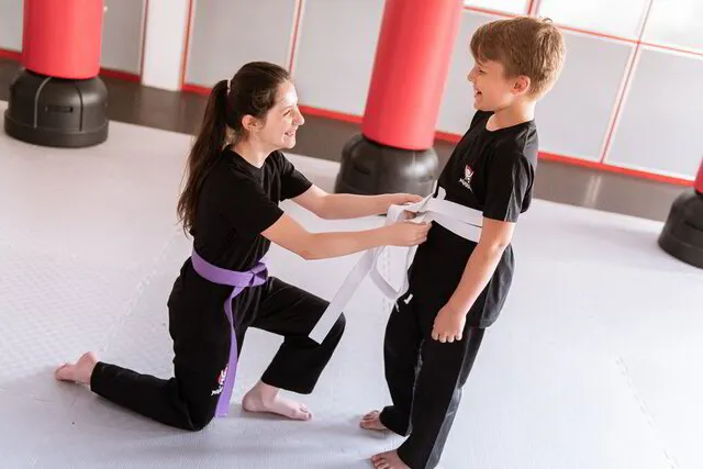 Ein fortgeschrittenes Karate Mädchen mit violettem Gürtel hilft einem neuen Schüler seinen weißen Gürtel zu binden, beide lächeln sich an, Unterstützung und neue Freundschaften werden bei Karate Geiger großgeschrieben