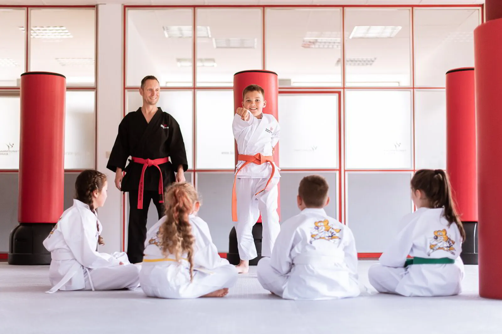 Mitarbeiter steht Kind zur Seite während es vor der Gruppe eine Karate Technik vormacht um sein Selbstbewusstsein zu stärken