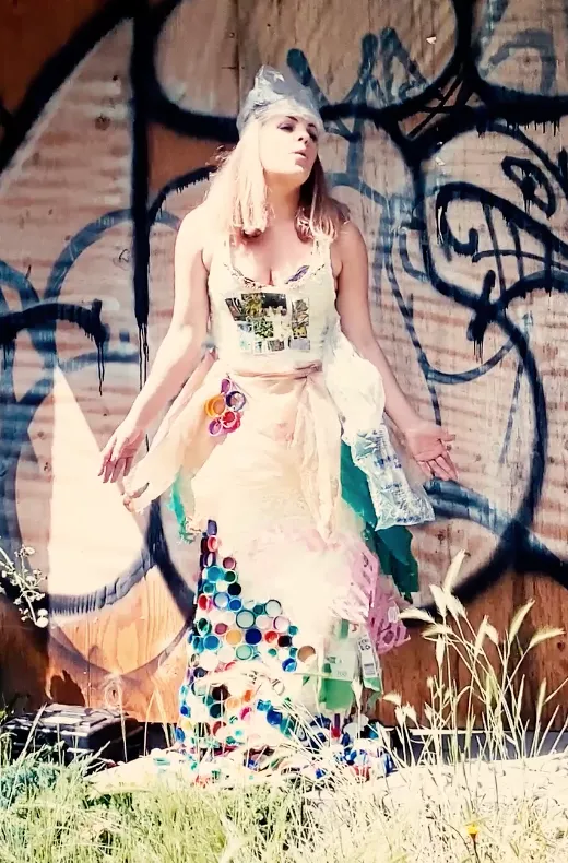 Still shot from Abby London's 'Anxiety' music video, showcasing the political anti-pop singer standing in front of a graffiti wall, wearing a dress crafted from recycled trash, encapsulating her creative persona.