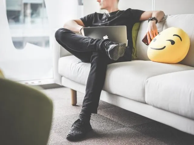 A man sitting on a couch with with electronics in lap and hand.